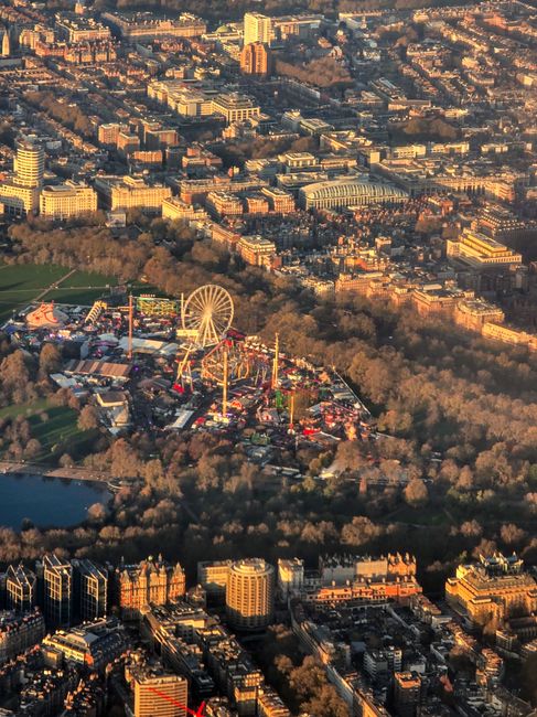 An aerial view of central London showing densely packed historic and modern buildings, with a large green park in the foreground containing an amusement park featuring a prominent Ferris wheel and sev